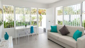 A bright living room with a white sofa, teal cushions, a glass dining table, and clear chairs.