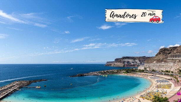 A coastal beach scene with a clear blue sky, a large sandy beach, and a rocky cliff in the background.