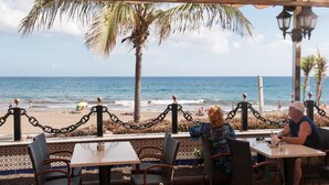 An outdoor dining area with tables and chairs, overlooking a beach with palm trees and the ocean.