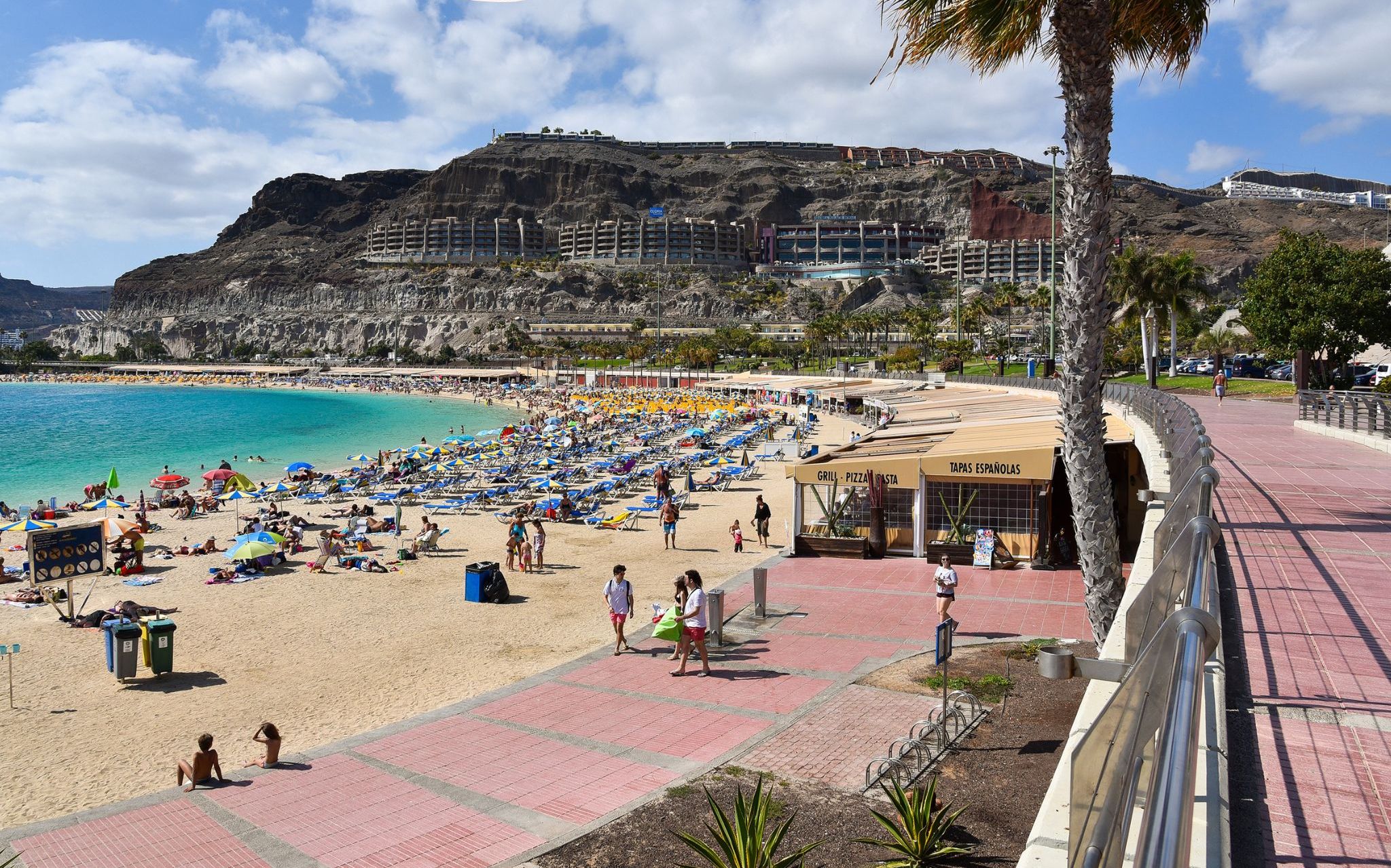 A beach with a rocky cliff in the background, a paved walkway, and a restaurant named 'GRILL - PIZZA PASTA'.