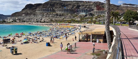 A beach with a rocky cliff in the background, a paved walkway, and a restaurant named 'GRILL - PIZZA PASTA'.