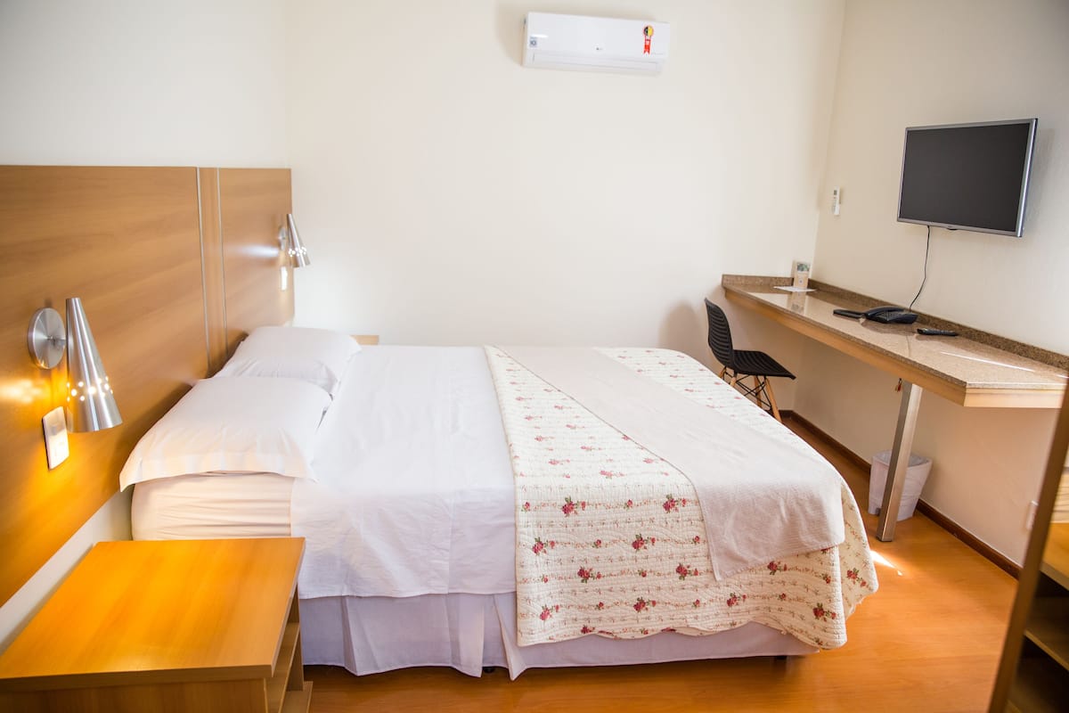 Une chambre d’hôtel équipée d’un lit, d’une table de chevet, d’un bureau, d’une chaise, d’une télévision et d’un appareil de climatisation.