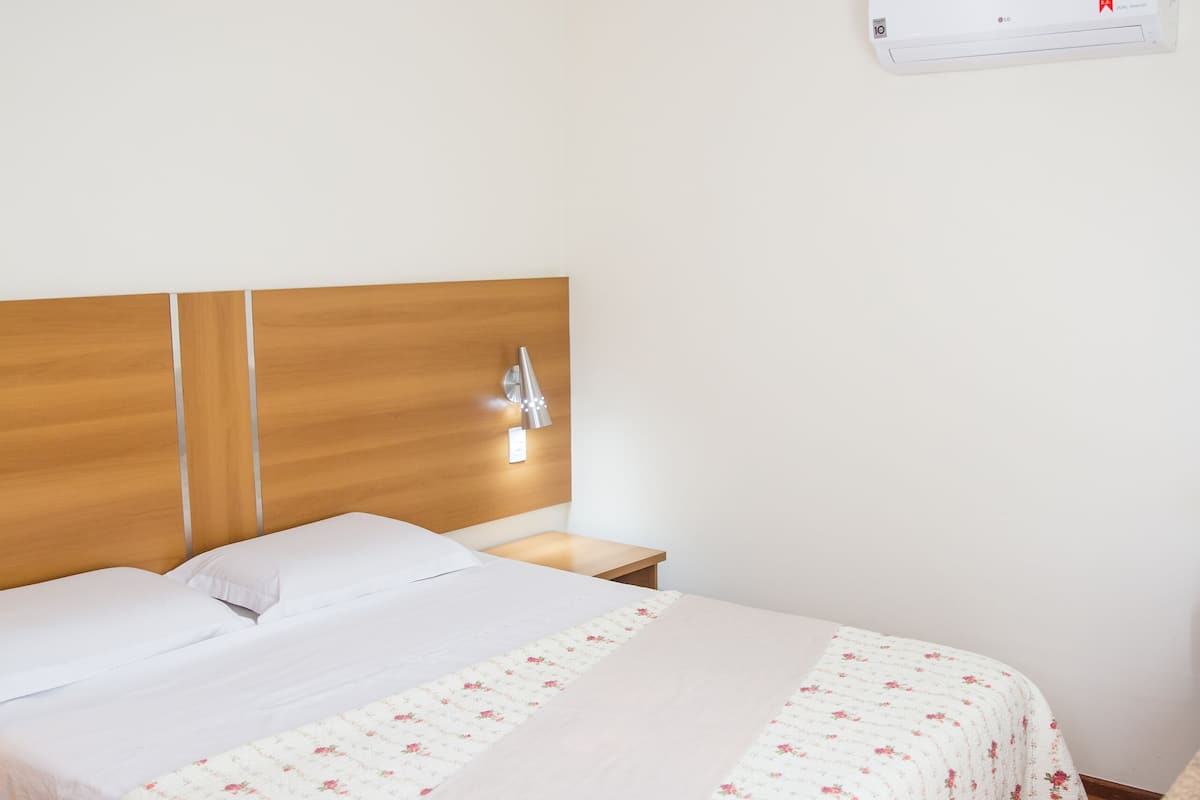 Une chambre d’hôtel avec une tête de lit en bois, un lit simple recouvert d’une couvre-lit à motifs floraux, une lampe de chevet, une table de chevet et un climatiseur fixé au mur.