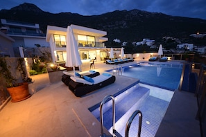 A modern outdoor pool area with lounge chairs and umbrellas, surrounded by a mountain backdrop. - Contemporary Villa, amazing sea views, heated infinity pool, daily maid service (Kiziltas, Kalkan)