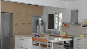 A modern kitchen with a stainless steel refrigerator, white cabinets, a large window, and a dining area with two chairs. - 'KATIKIES 22' Luxury Detached Pissouri Beach Properties, Great Relaxing Location (Pissouri)