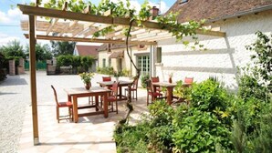 A covered patio with wooden tables and chairs, a white building with a tiled roof, and a gravel path. - Maison Cerviña (Germigny-des-Pres)
