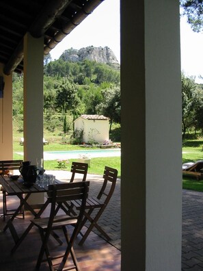 Une terrasse couverte, avec une table et des chaises, donnant sur un jardin et une montagne.