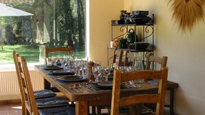 A dining table set for four with chairs, a metal shelf with kitchenware, and a view of greenery outside. - Villa with Pool, Privacy and Magnificent View (Barbentane)