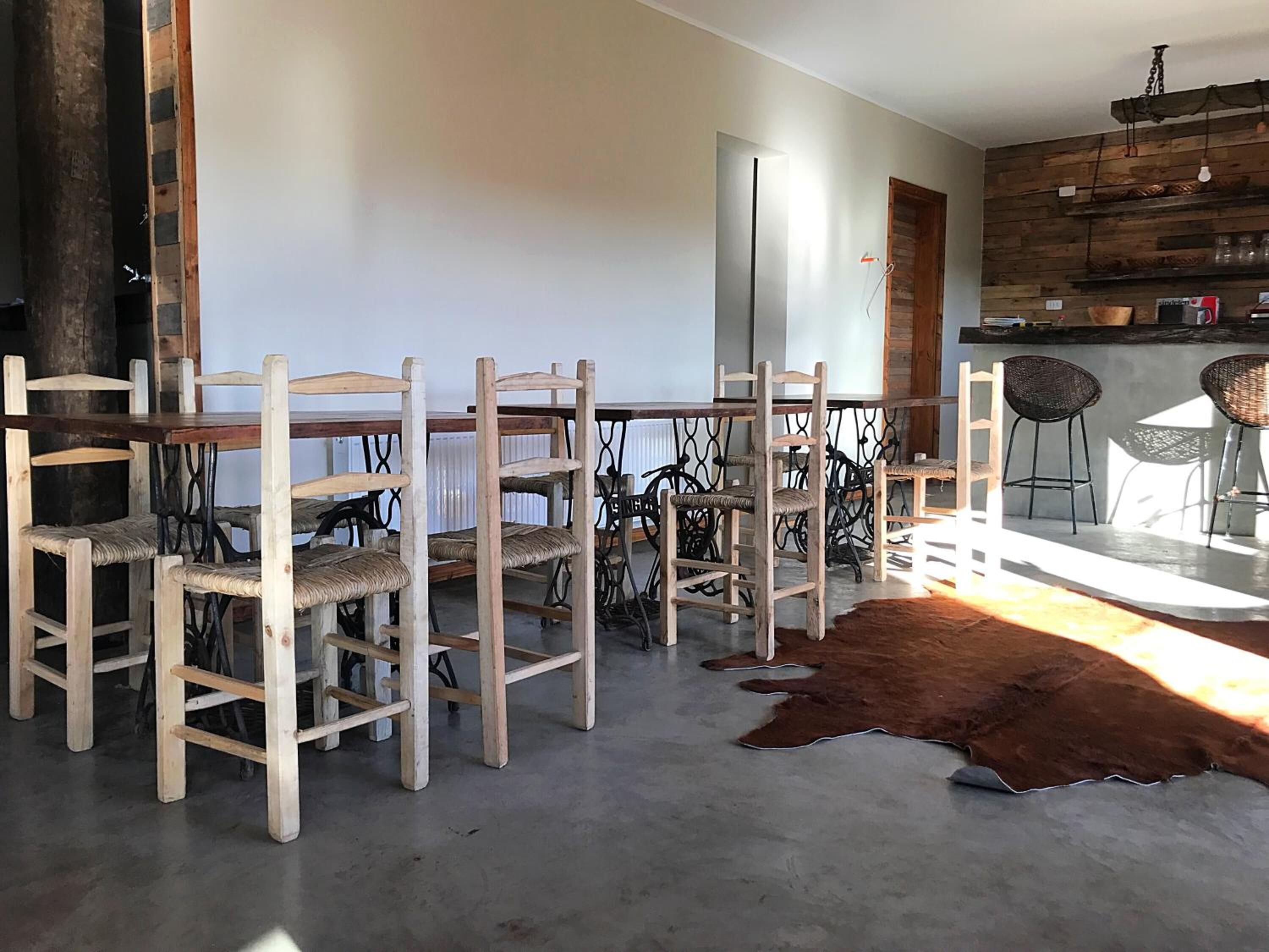 Un comedor con sillas de madera y una mesa, un tapete de cuero de vaca, y una zona de cocina con taburetes de bar y estantes.
