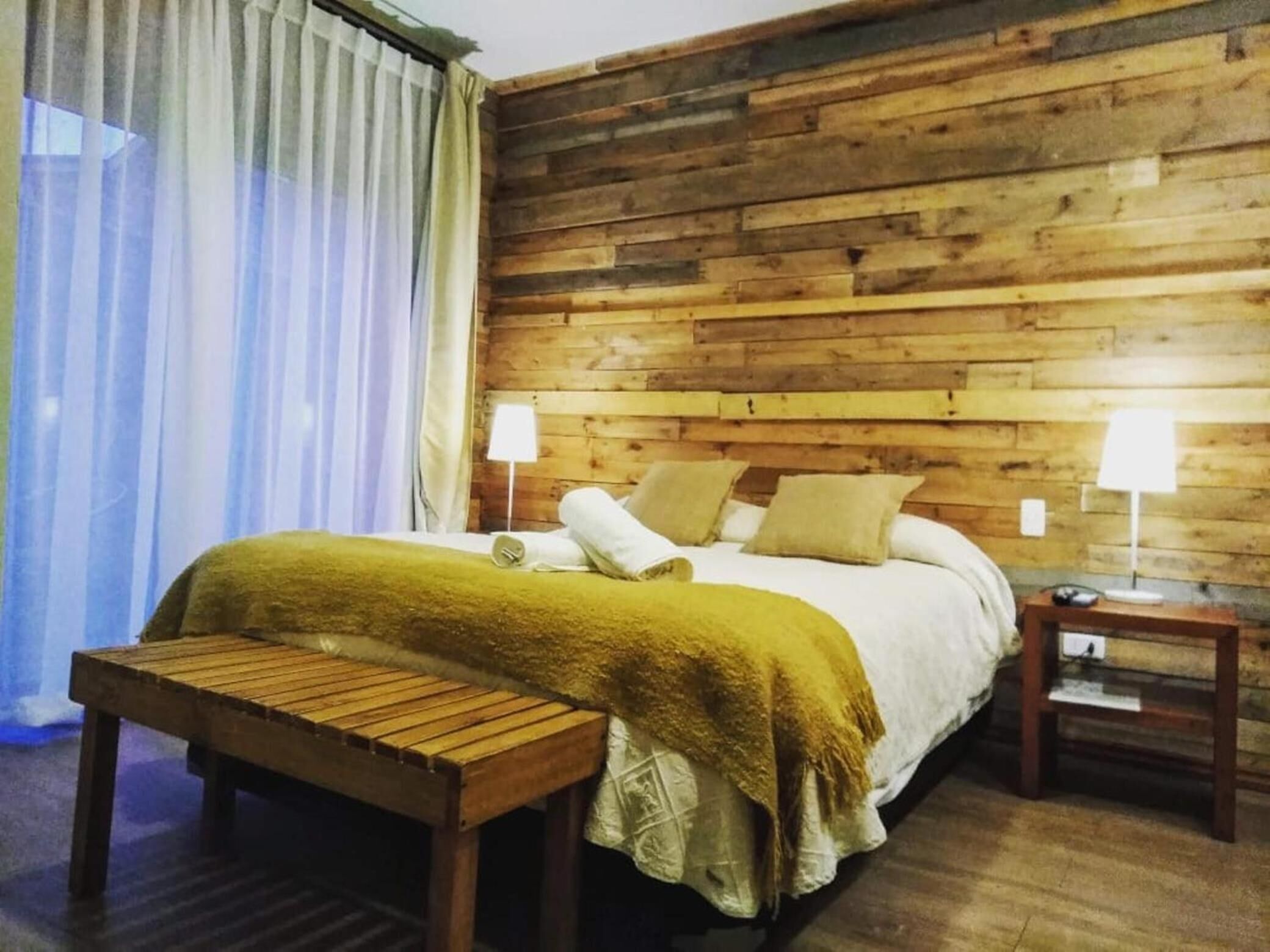 Una habitación con paneles de madera, una cama, dos mesitas de noche, un banco y una ventana con cortinas.
