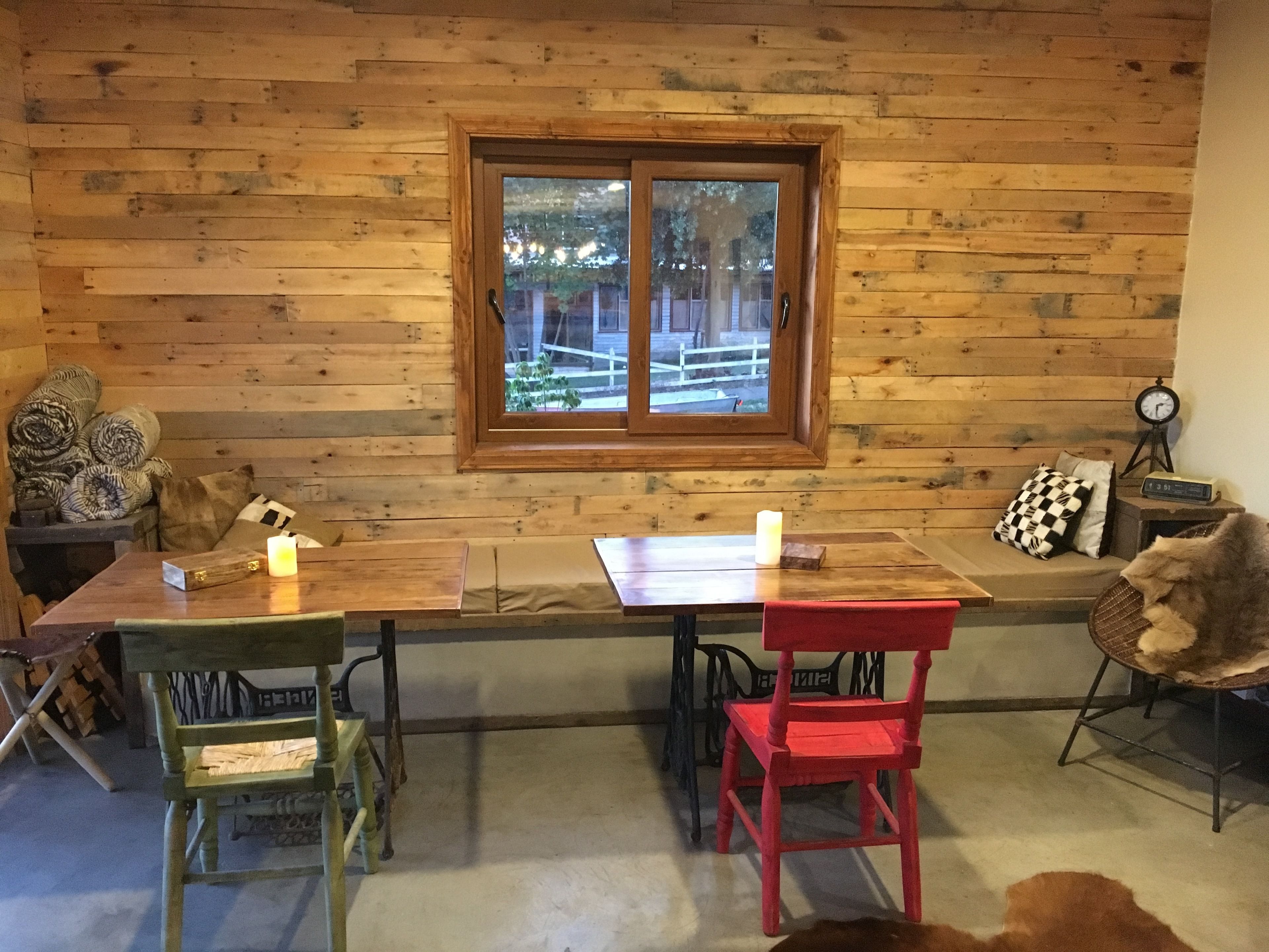 A wooden cabin interior with a dining table, two chairs, a bench, and a window overlooking a fenced yard.
