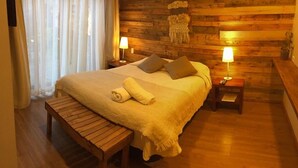 A cozy bedroom with a wooden headboard, a large bed with pillows, a bedside table with a lamp, and a bench. - Hotel 381 (Panguipulli)