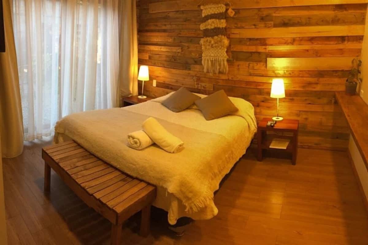 A cozy bedroom with a wooden headboard, a large bed with pillows, a bedside table with a lamp, and a bench.