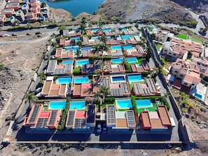 An aerial view of a residential area with multiple houses, each with a swimming pool and solar panels.
