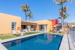 A modern outdoor pool area with lounge chairs, a barbecue, and palm trees.