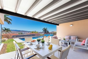 A covered patio with a pool view, white furniture, and a table set for dining.