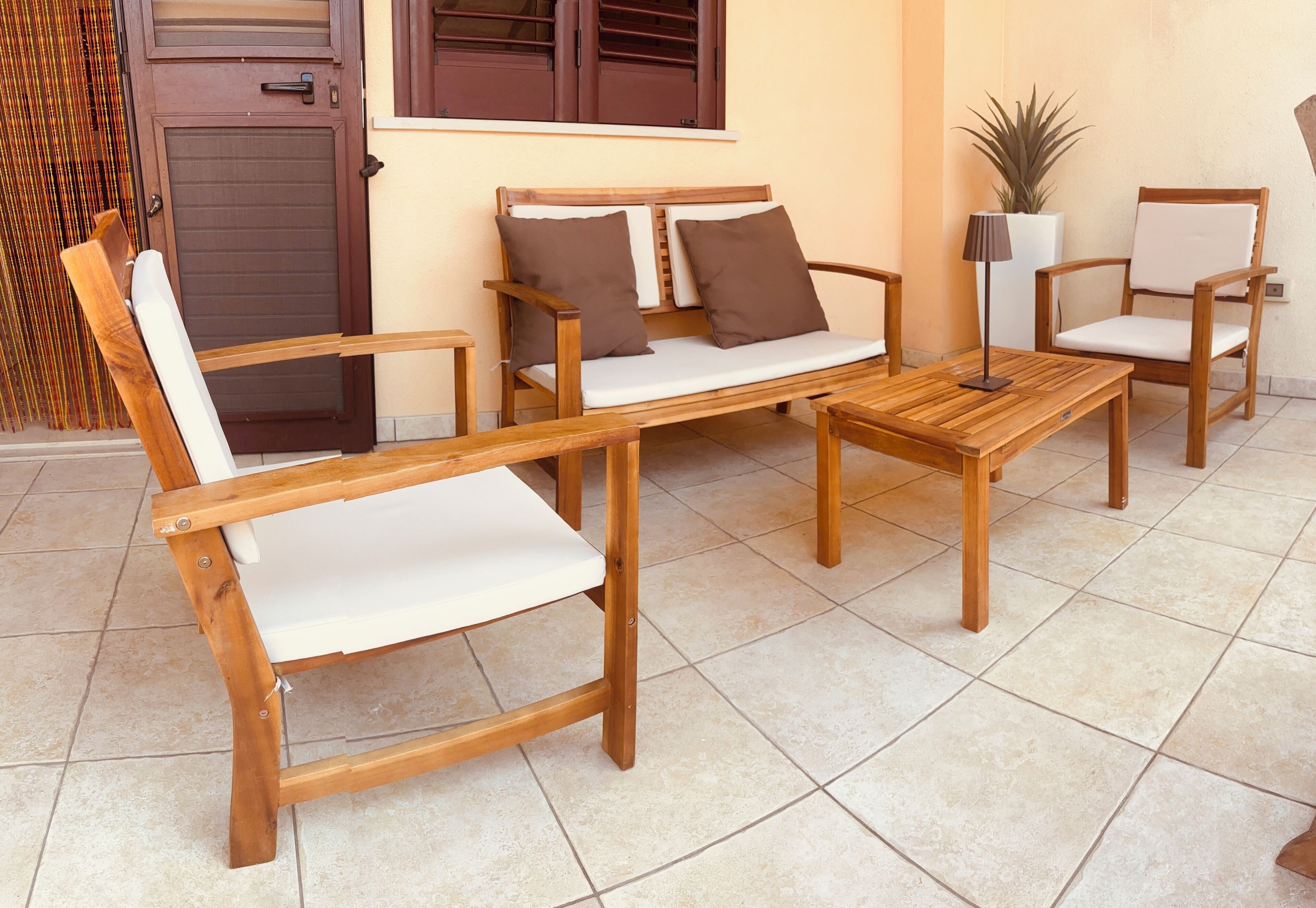 A wooden sofa with cushions, a wooden table, and a chair in a sunlit outdoor area with tiled flooring.