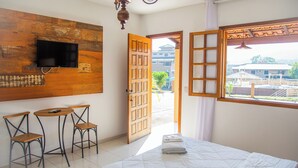 Habitación de hotel con cama, televisor montado en una pared de madera, dos sillas, una mesita y vistas al exterior a través de una puerta abierta.