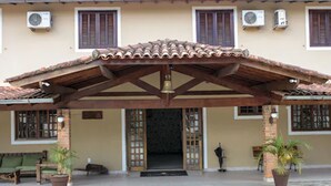 Un edificio con techo de tejas, una pérgola de madera y una puerta con celosía decorativa. Hay plantas en macetas y un banco afuera.
