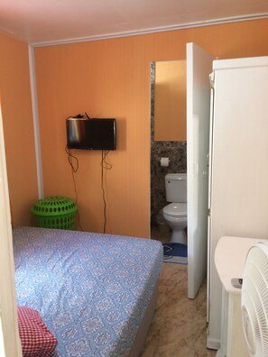 Una habitación pequeña con una cama, televisor montado en la pared, un inodoro y un refrigerador.