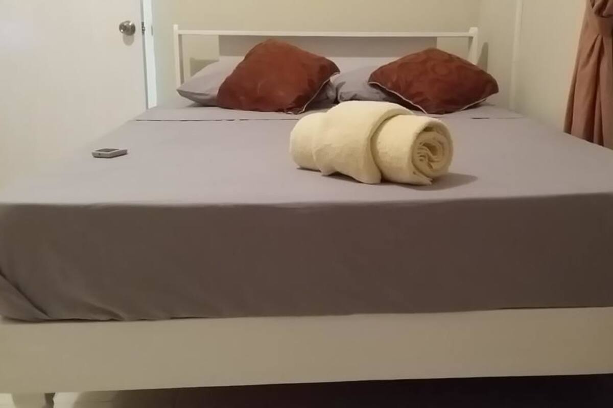 Una cama con una colcha gris, dos almohadas marrones y una toalla blanca enrollada.