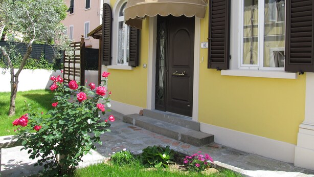 Ein gelbes Gebäude mit brauner Tür und Fensterläden, einer kleinen Veranda mit beiger Markise und einem Garten mit rosa Blumen.