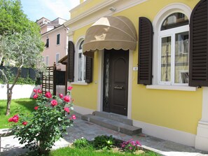 A yellow building with a brown door and shuttered windows, a small porch with a beige awning, and a garden with pink flowers. - In the heart of beautiful Tuscany, Spa, Culture and Good Food (Monsummano Terme)