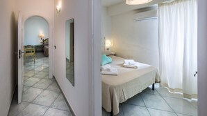 Habitación de hotel con cama, mesita de noche, lámpara y vista al pasillo.