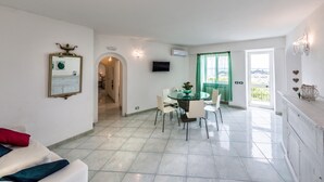 Una sala de estar moderna con un sofá blanco, un comedor con mesa de vidrio y vistas a la ciudad desde un balcón.