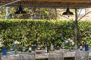 Un área de comedor al aire libre con mesas dispuestas para una comida, rodeada por un denso seto verde.