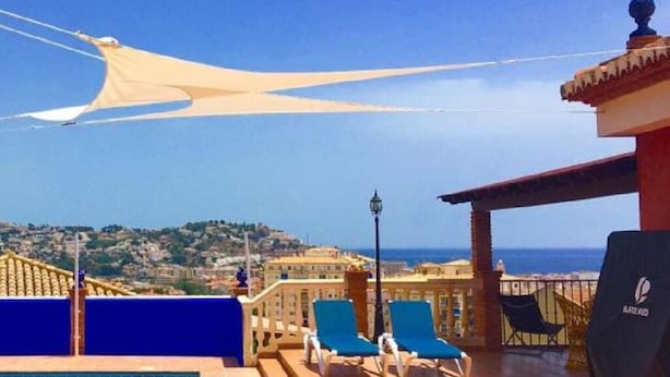 Zona de piscina en la azotea con vista al mar, dos sillones azules y una pérgola con toldo.