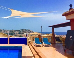 A rooftop pool area with a view of the sea, two blue lounge chairs, and a pergola with a canopy.