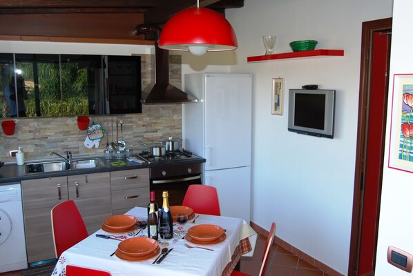 A kitchen with a dining table set for two, a red pendant light, and a refrigerator.