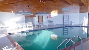 An indoor swimming pool with clear water, surrounded by white lounge chairs and striped umbrellas. - AmericInn by Wyndham Madison SD (Madison)