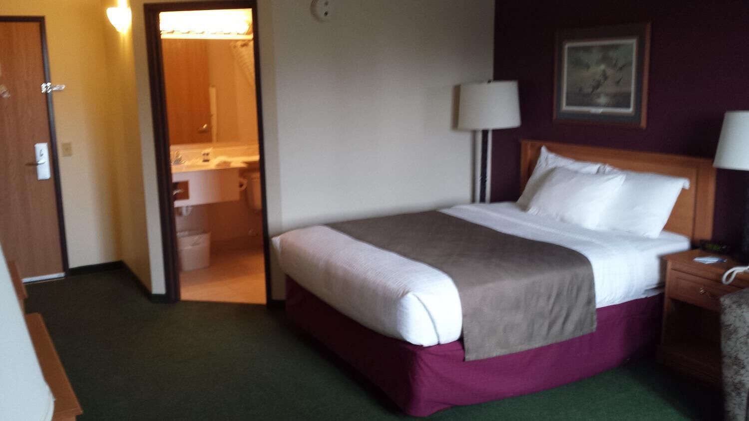 A hotel room with a bed, a nightstand, a lamp, and a bathroom visible through an open door.