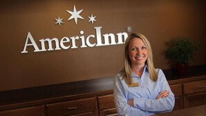 A person standing in front of a wall with the American Inn logo, a wooden counter, and a plant. - AmericInn by Wyndham Madison SD (Madison)
