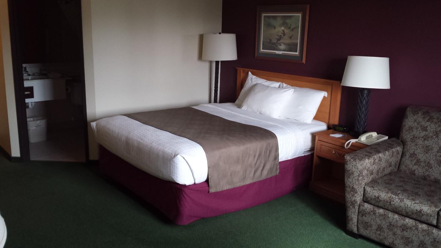 Una habitación de hotel con cama, dos lámparas de noche, una silla y un teléfono en la mesita de noche.