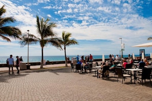 An outdoor café with tables and chairs, palm trees, and a view of the sea.