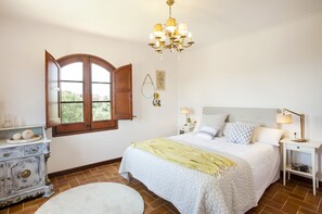 Une chambre à coucher avec un lit, des tables de chevet, un lustre, une commode et une fenêtre dont les volets sont ouverts.