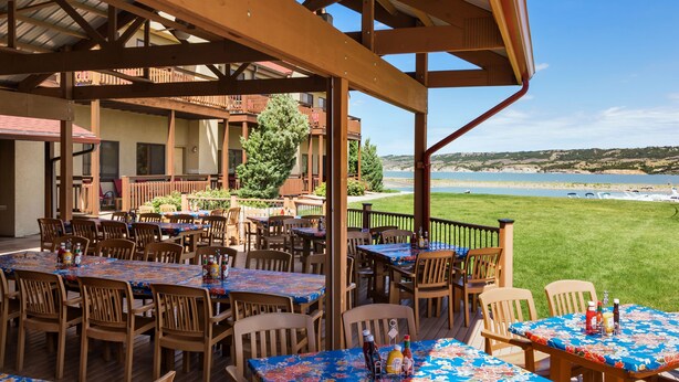 An outdoor patio with multiple wooden tables and chairs, a view of the beach, and a building with balconies in the background.