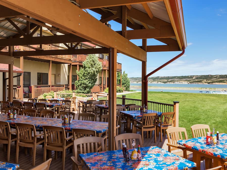 An outdoor patio with multiple wooden tables and chairs, a view of the beach, and a building with balconies in the background.