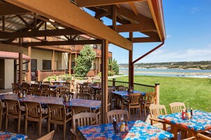 Un patio al aire libre con varias mesas y sillas de madera, vista a la playa y un edificio con balcones al fondo.