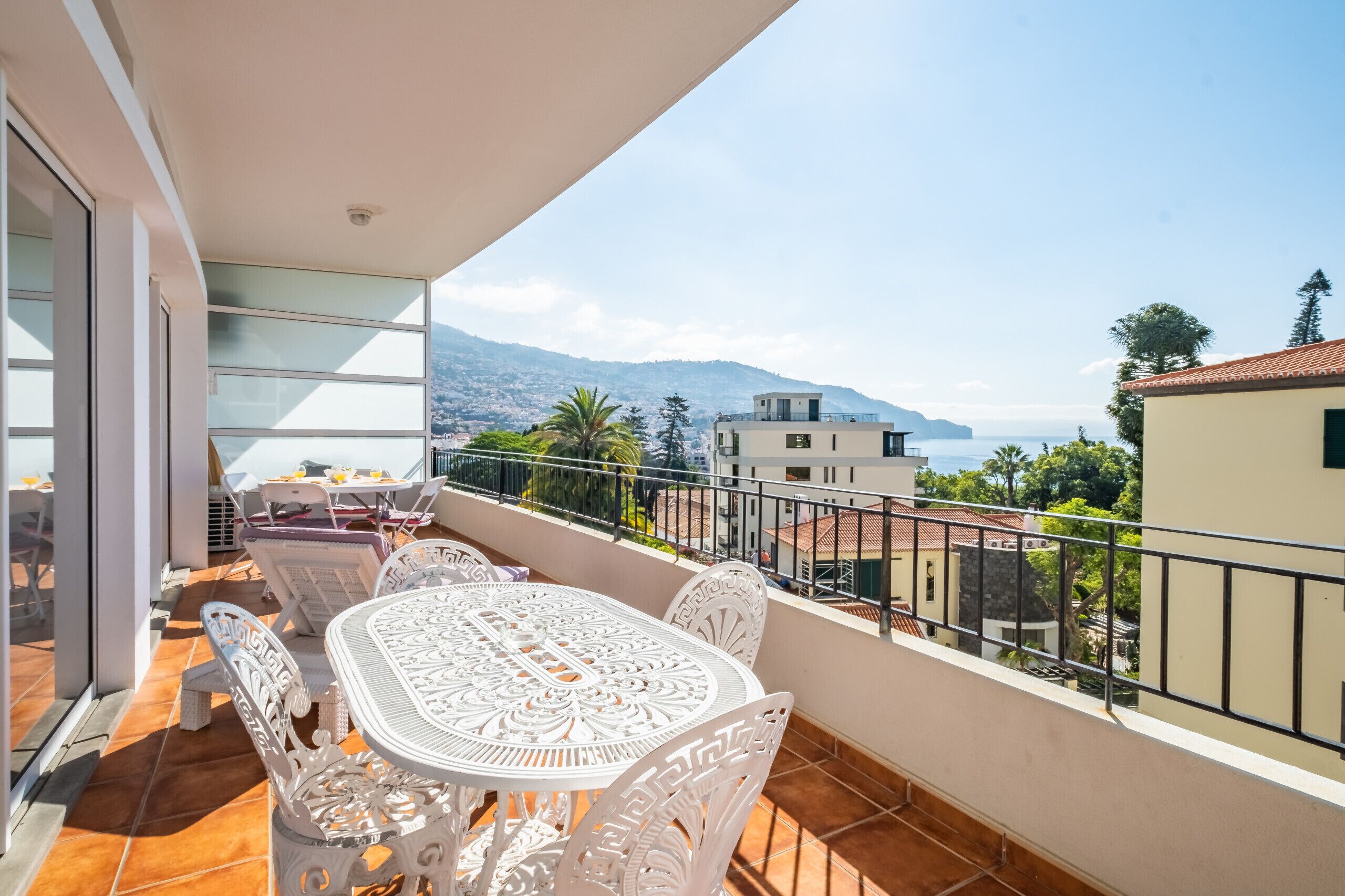 A balcony with a table and chairs, overlooking a coastal view with buildings and mountains.