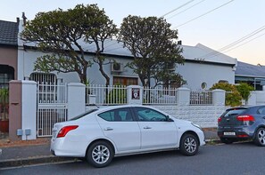 A white car parked in front of a white building with a gated entrance, trees, and a clear sky.