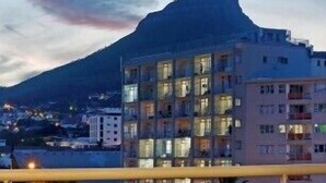 Un balcón con mesa y sillas, una botella de vino tinto y una copa. Al fondo, se ve una montaña y un edificio de varios pisos.