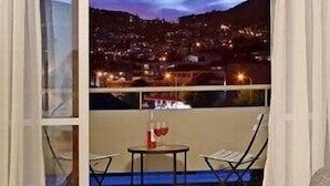 Una habitación con un amplio ventanal corredera que da a un balcón con vistas a un paisaje urbano al atardecer.
