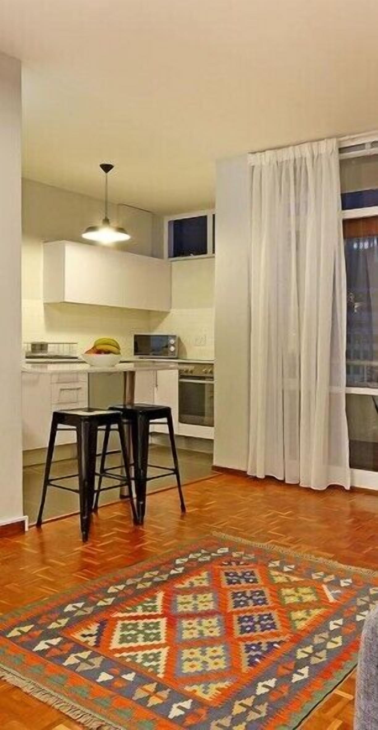 A compact kitchen with white cabinets, a microwave, and a fruit bowl on the counter. There are two black stools and a patterned rug on the wooden floor.