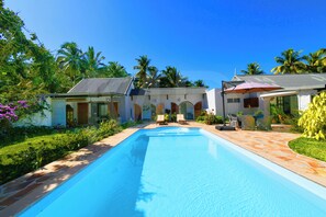 A swimming pool with lounge chairs and a covered patio area, surrounded by lush greenery and palm trees.