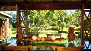 Un área de comedor al aire libre con una mesa de vidrio, sillas de madera y un banco de madera. Hay piñas y naranjas sobre la mesa.