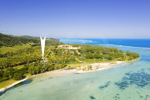 An aerial view of a coastal area with a prominent white wind turbine, lush greenery, and clear blue waters.